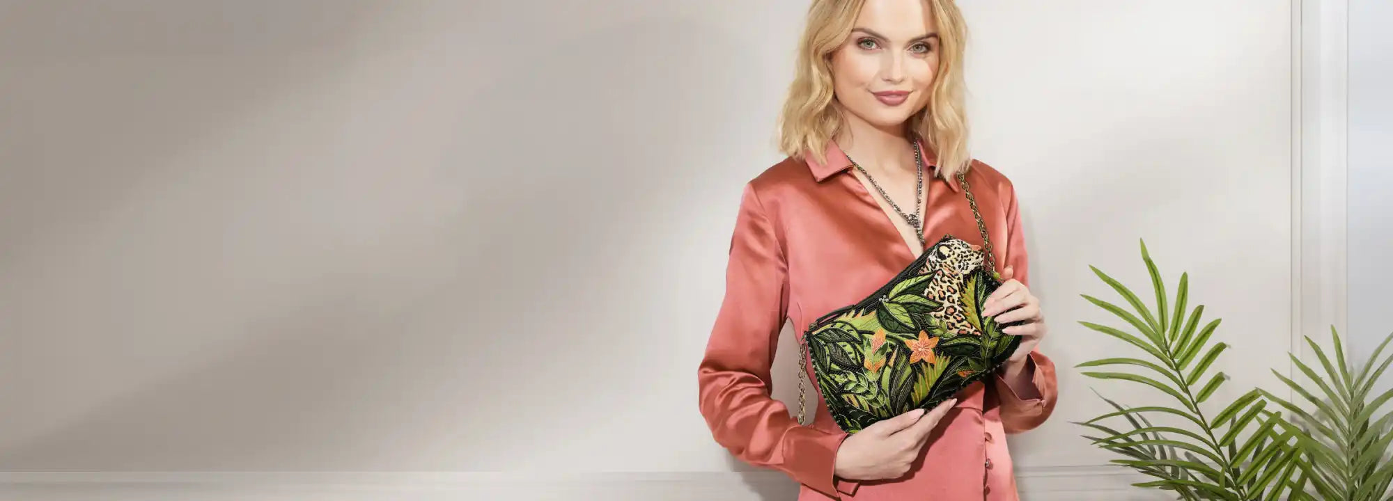 A blonde woman in a pink satin shirt holding a rectangular clutch bag with green tropical leaf and orange flower embroidery.