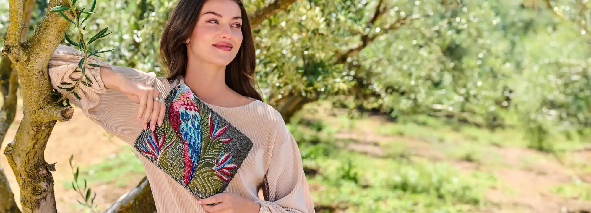 A woman modeling a long-sleeved, off-the-shoulder top and holding a colorful clutch bag with a parrot design.